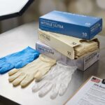 Three types of disposable gloves—blue nitrile, natural latex, and clear vinyl—fanned out on a workbench next to a PPE checklist and an open glove box